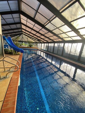 An indoor swimming pool with a blue slide, tiled flooring, and a covered roof structure.