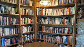 A wooden table surrounded by chairs in a room with tall bookshelves filled with books. - Charming Log Cabin Retreat with Flower Garden (McKenzie)