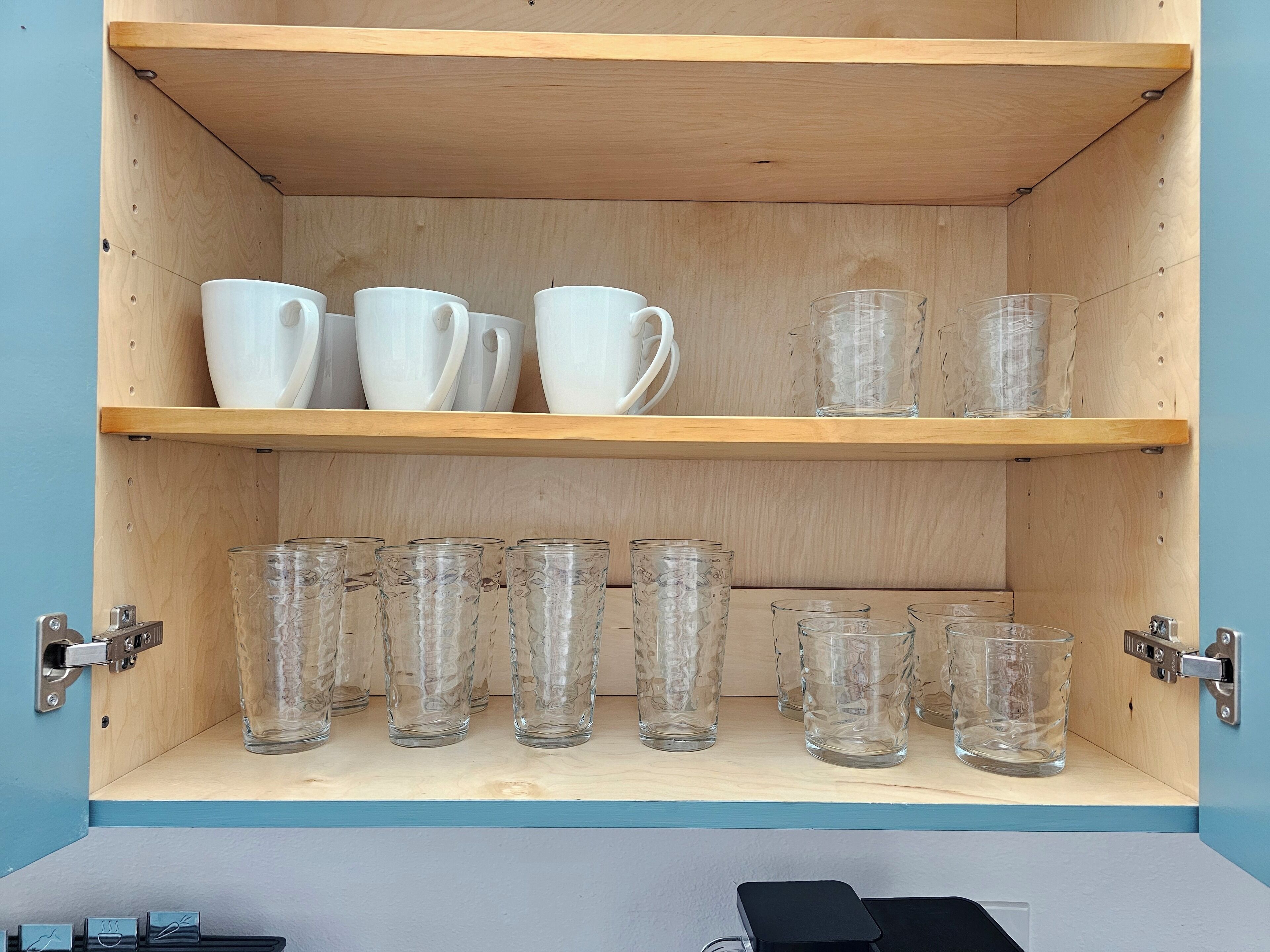 Eine Holzschrank mit zwei Einlegeböden, auf denen weiße Tassen und klare Glasbecher stehen.