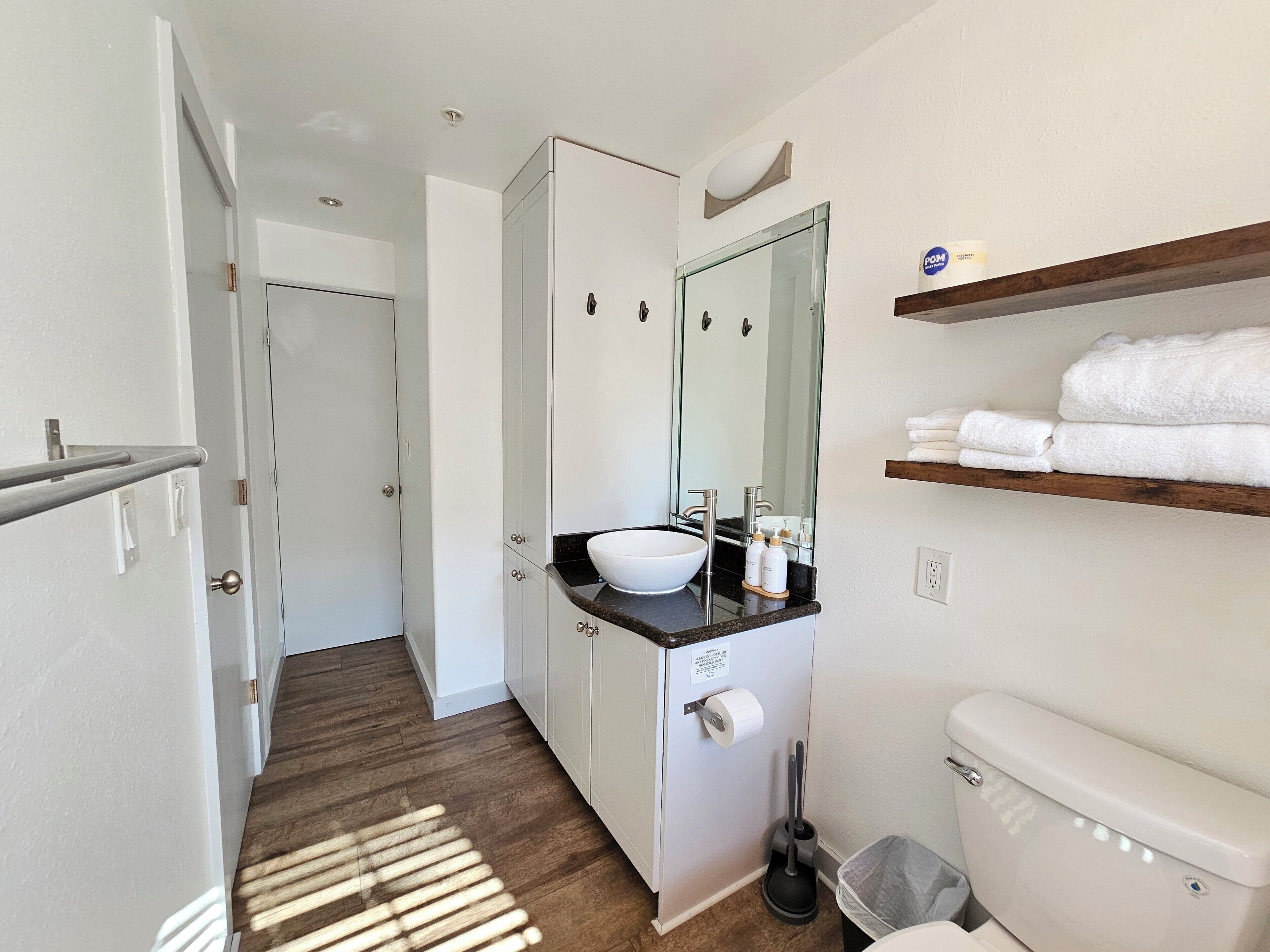 A modern bathroom with a white sink, mirror, and toilet. There is a wooden shelf with towels and a broom.