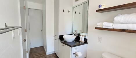 A modern bathroom with a white sink, mirror, and toilet. There is a wooden shelf with towels and a broom.