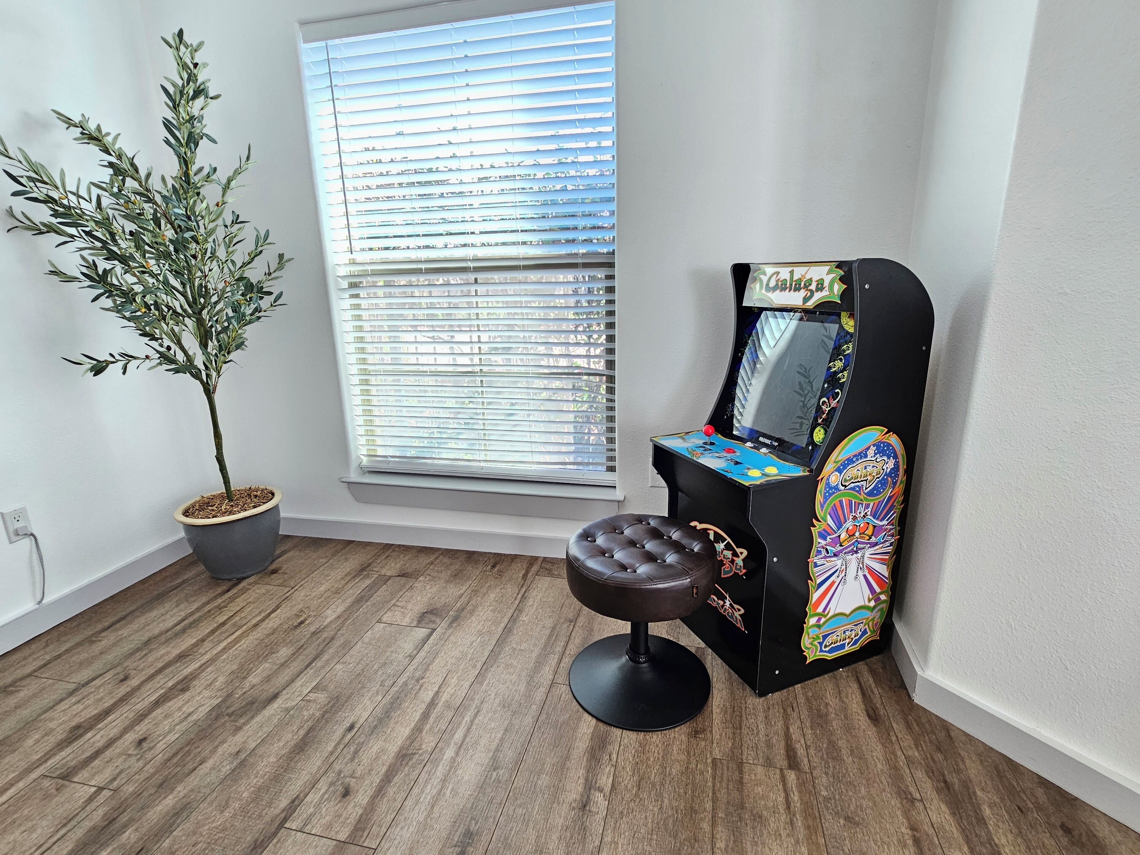 A room with a potted plant, a window with blinds, a small stool, and a colorful arcade machine.