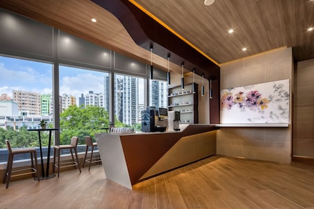 A modern reception area with a wooden reception desk, bar stools, a coffee machine, and a large window offering a city view.