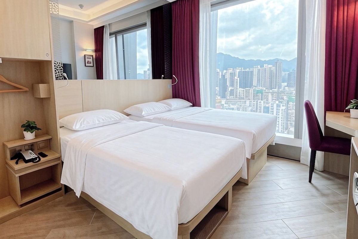 A hotel room with two beds, a desk, a chair, a small plant, and a view of the cityscape.
