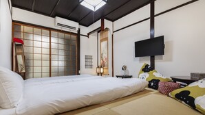 Een traditionele Japanse kamer met tatami vloeren, een futon bed, een verschuifbare shoji wand, een bevestigde televisie en een kleine tafel met een lamp.