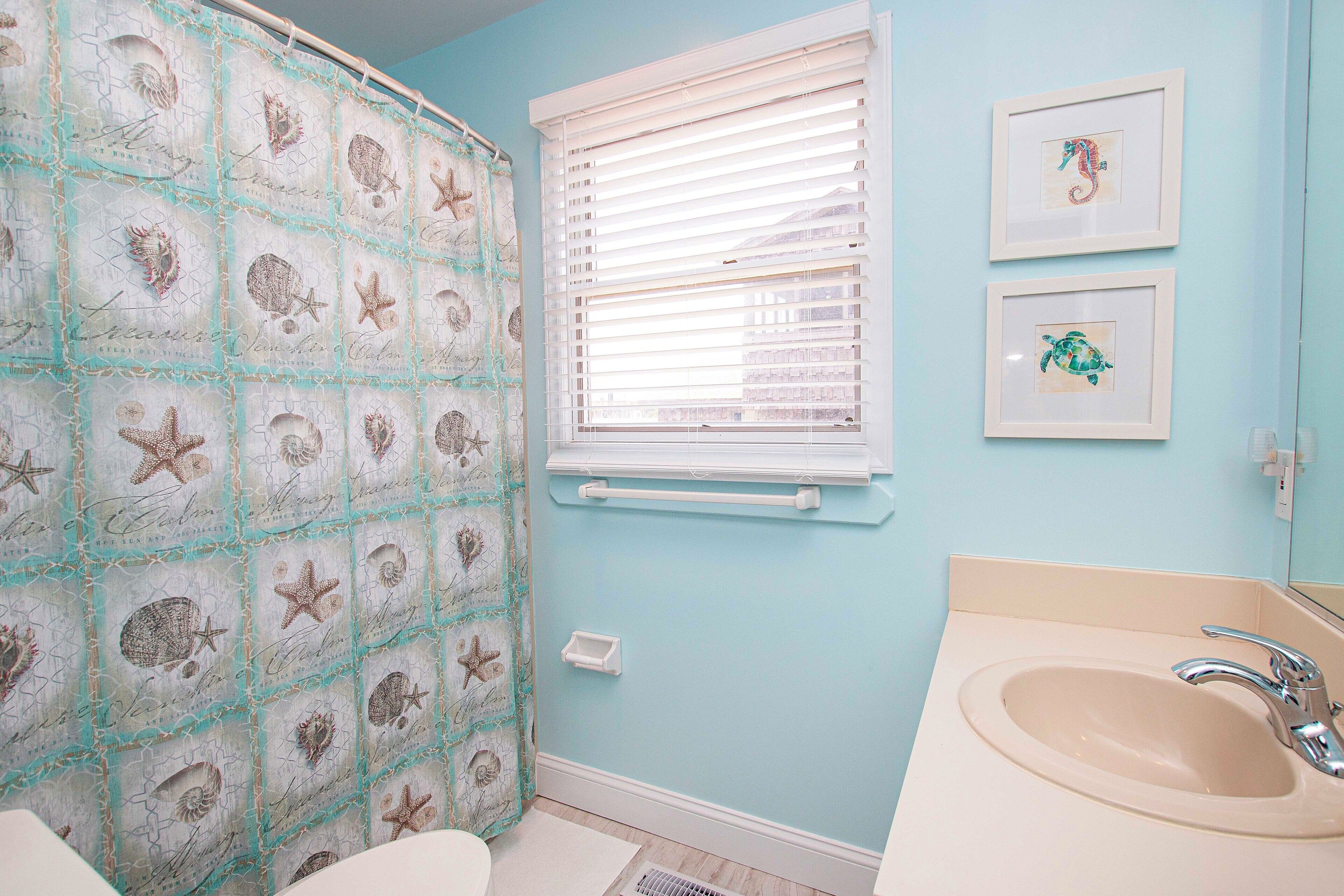 Een badkamer met een lichtblauwe wand, een douchegordijn met zeedieren, een raam met zonwering en een wastafel met spiegel.
