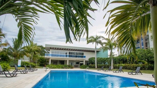 Una piscina con sillones de descanso, rodeada de palmeras y un edificio moderno con balcón de vidrio.