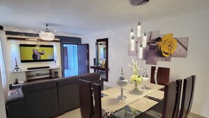Una sala moderna con sofá, televisor y comedor con mesa de vidrio y sillas oscuras.