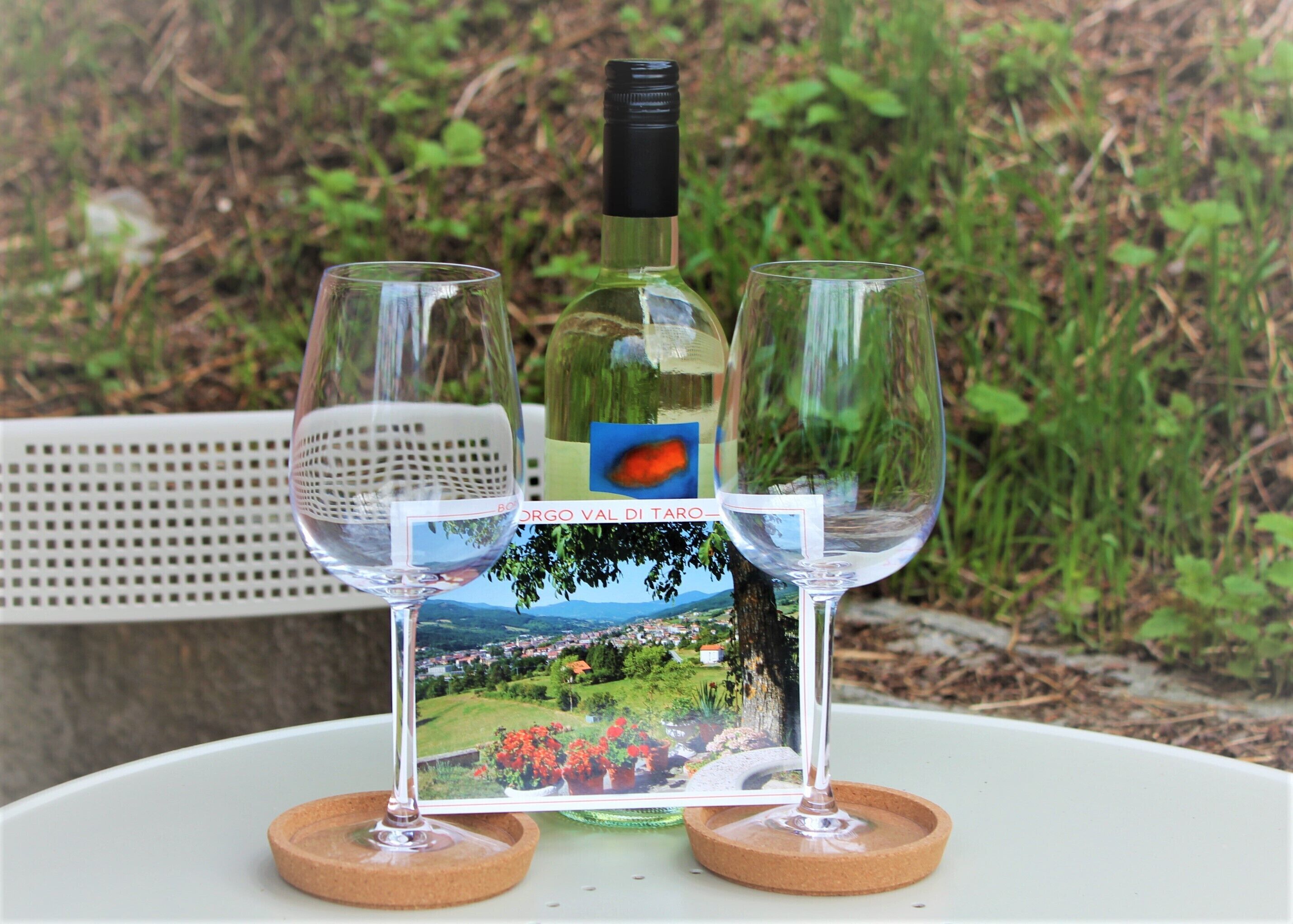Deux verres à vin sont posés sur une table, avec une bouteille de vin et une carte postale représentant un paysage pittoresque entre eux.