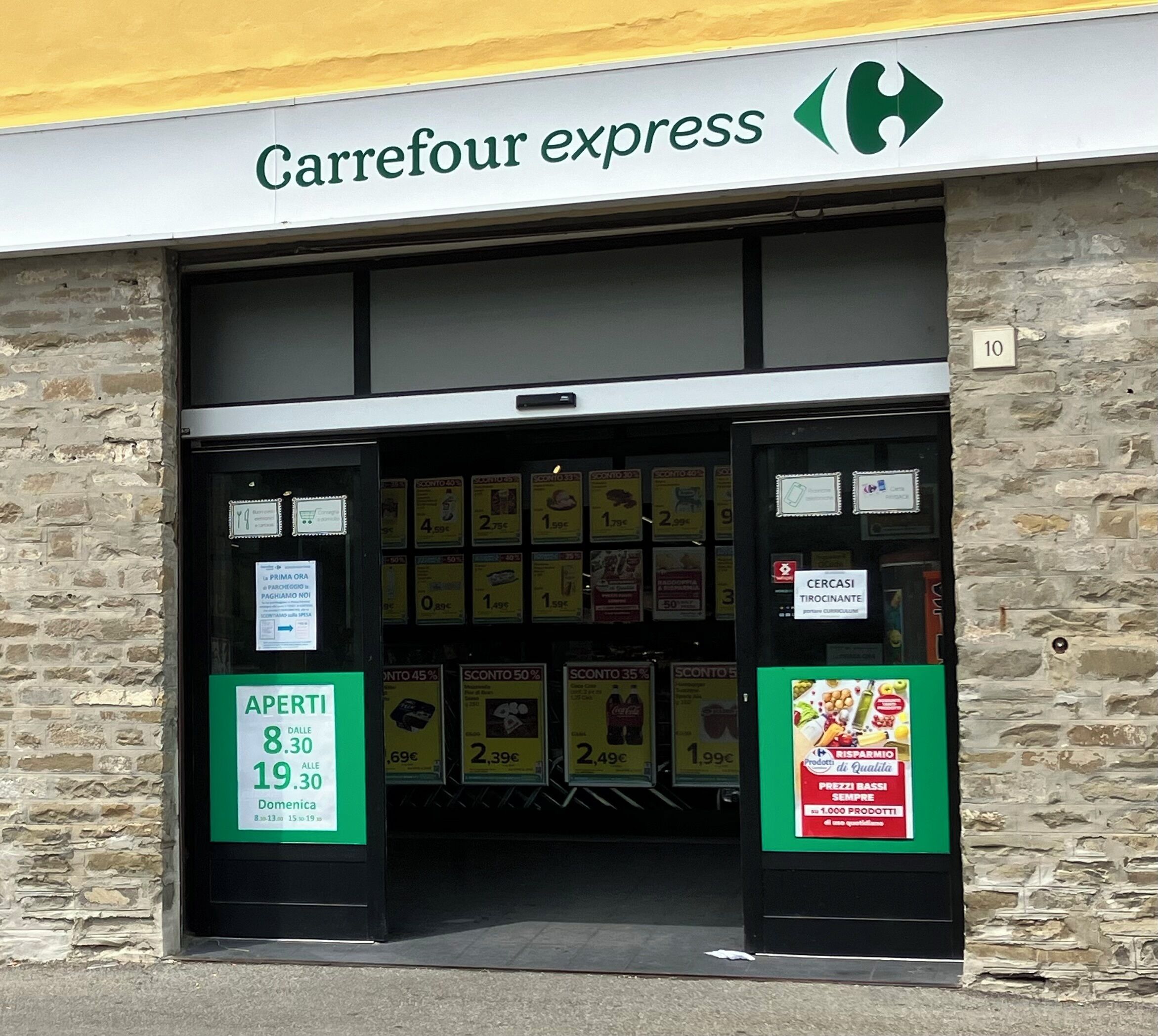 L’entrée d’un magasin Carrefour Express, avec une façade en pierre, des étiquettes de prix et des panneaux publicitaires.