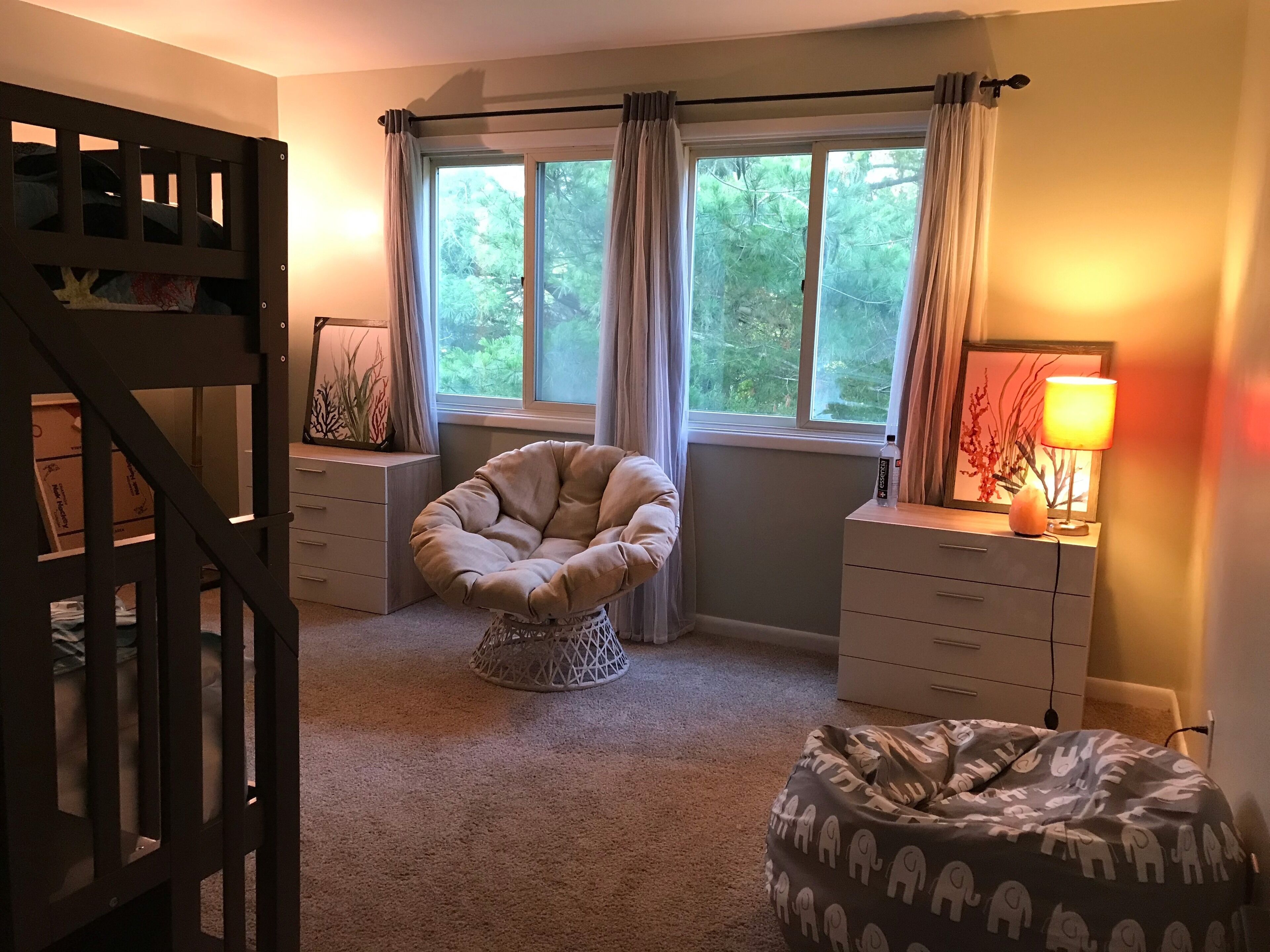 Ein Schlafzimmer mit Bett, Stuhl, Kommode, Lampe und einem Fenster mit Vorhängen.