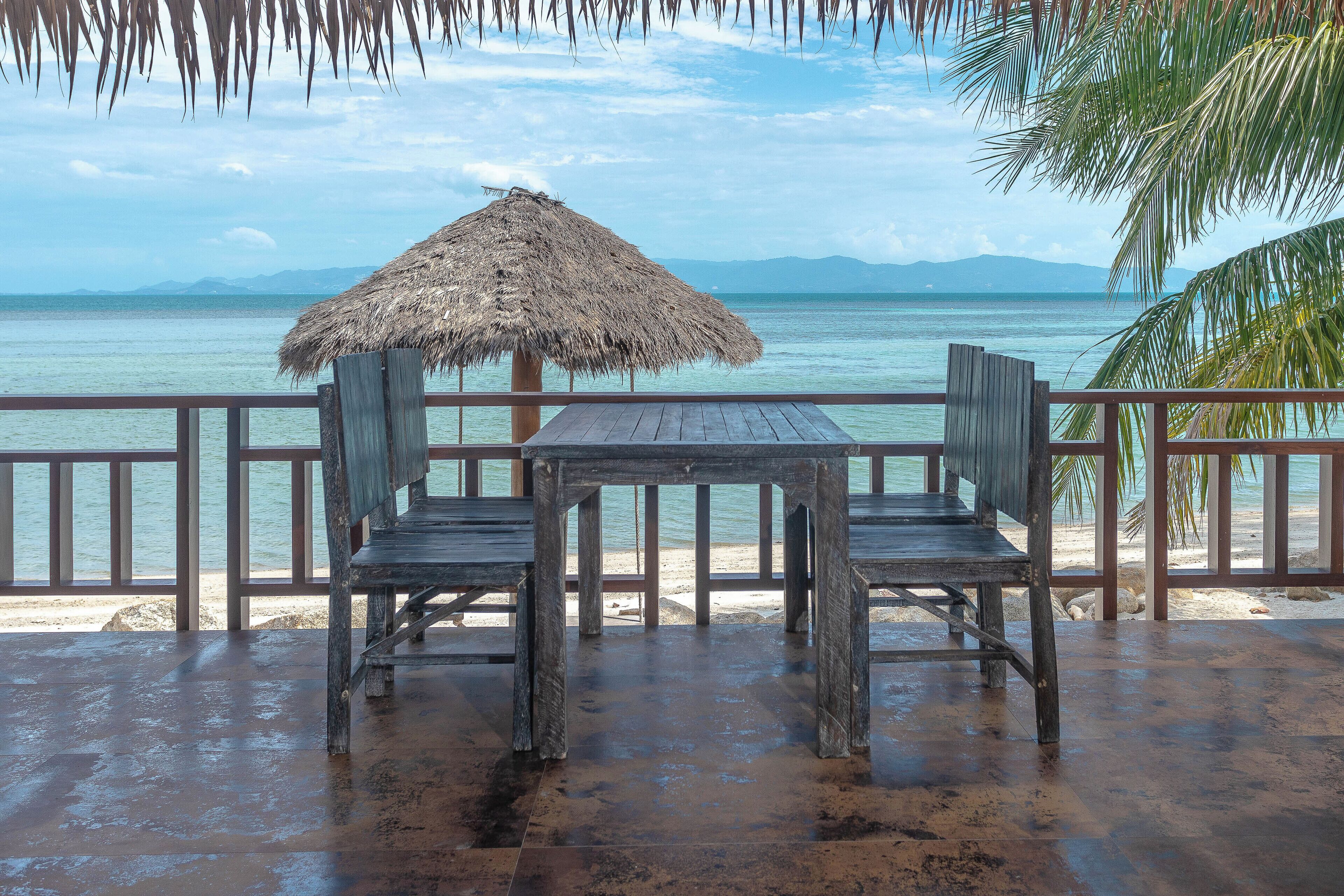 A wooden table and chairs under a thatched umbrella on a beachside deck with a view of the sea and mountains.