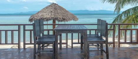 A wooden table and chairs under a thatched umbrella on a beachside deck with a view of the sea and mountains.