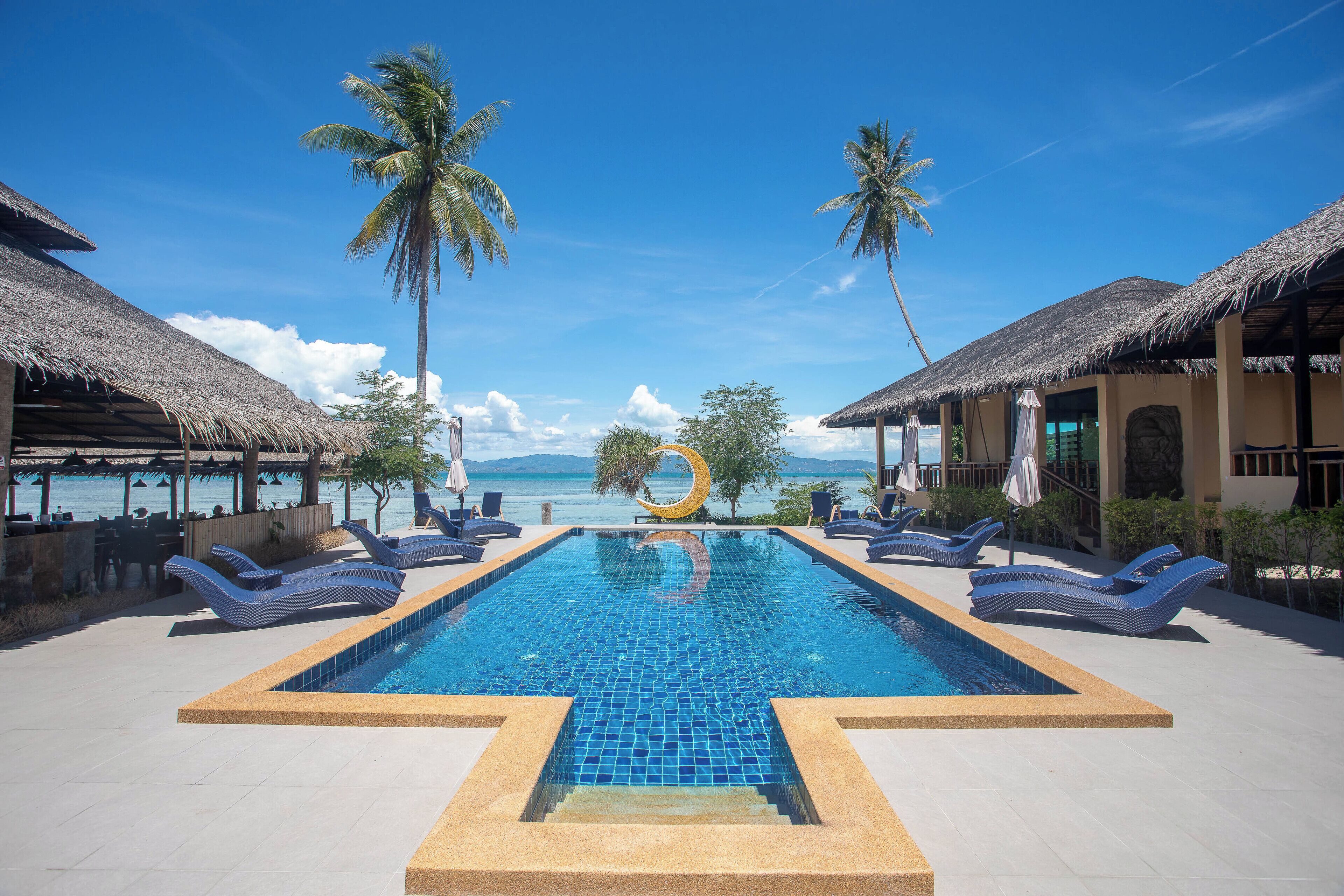 A pool with lounge chairs, palm trees, and a view of the ocean.
