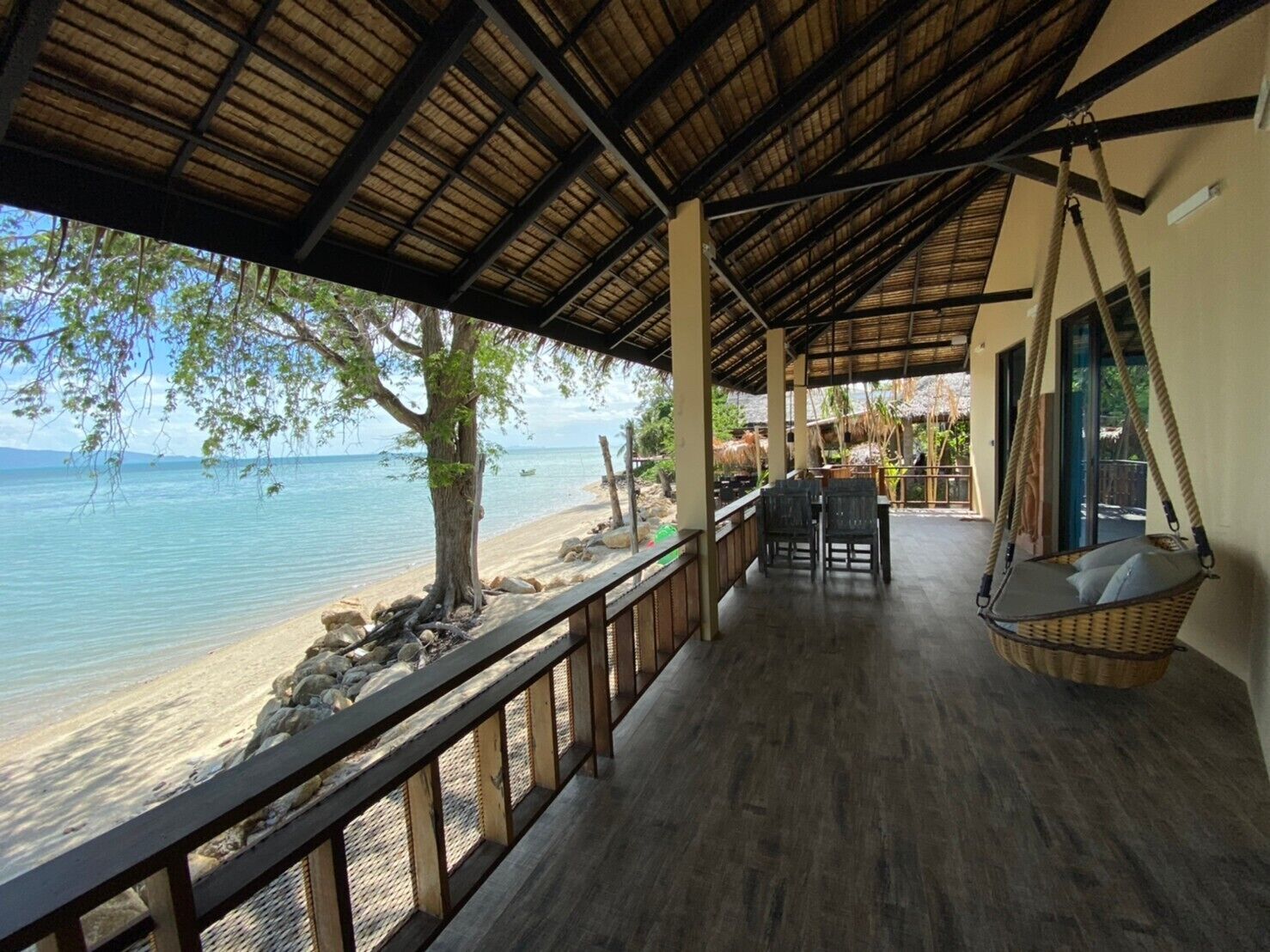 A covered outdoor area with a wooden deck, a hanging chair, and a view of the beach and sea.