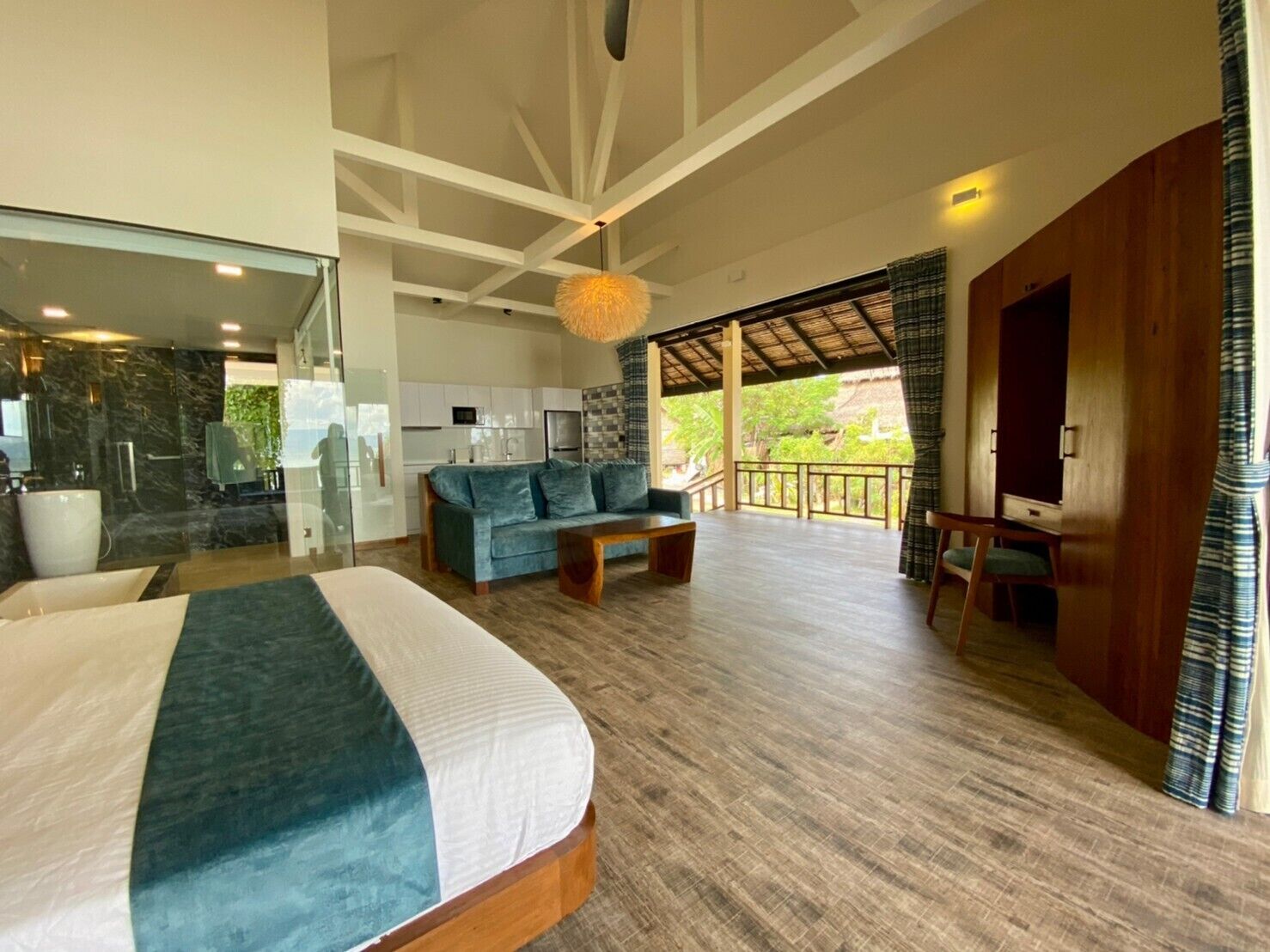 A spacious room with a large bed, a sofa, a dining table, and a balcony with a view of greenery.