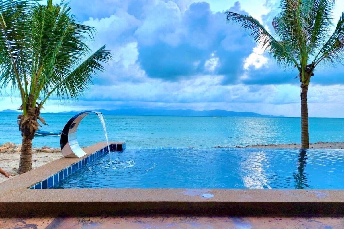 A swimming pool with a view of the sea and mountains, surrounded by palm trees.