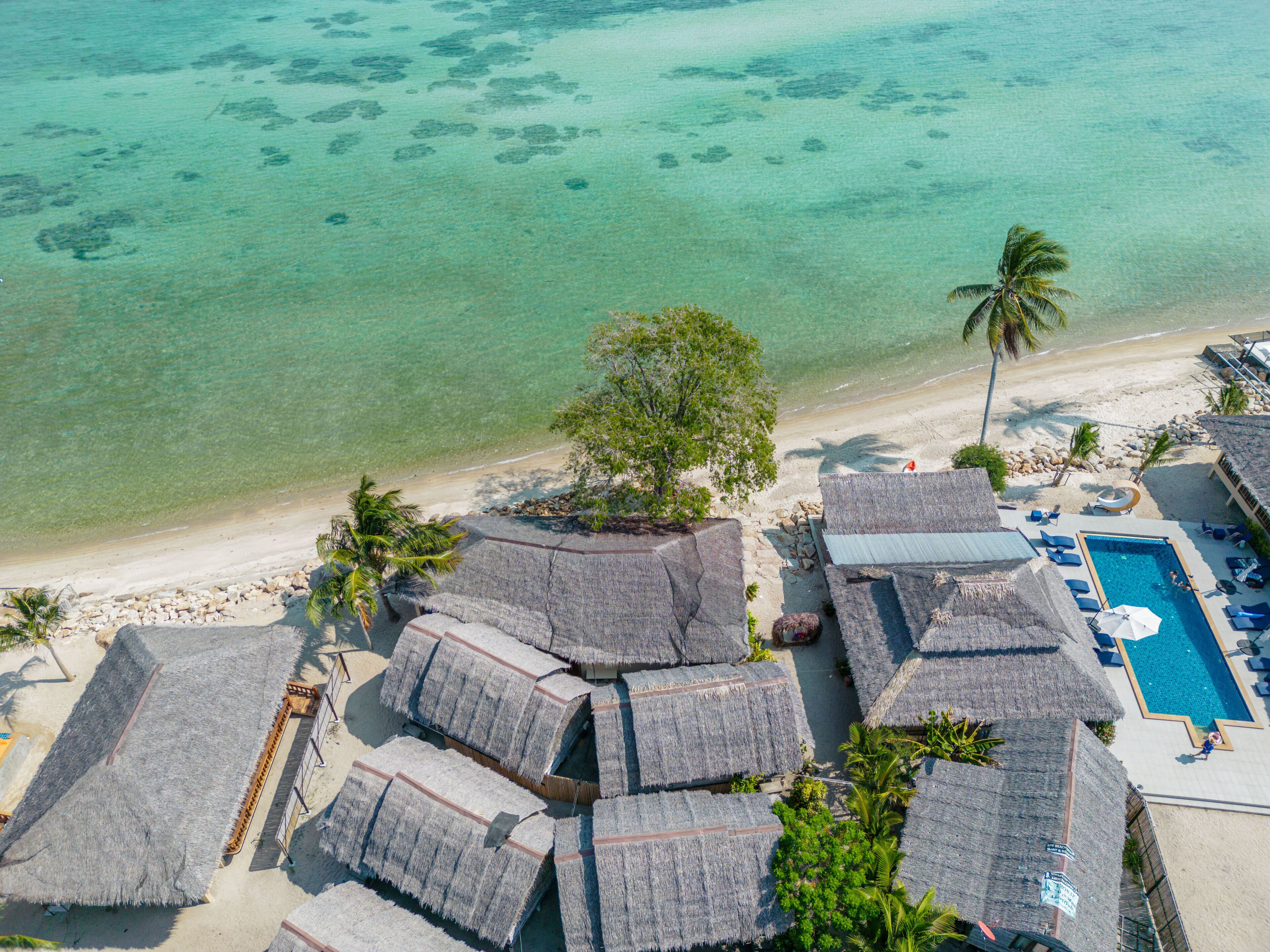 Vue aérienne d’un complexe hôtelier en bord de mer, avec des bungalows aux toits de chaume, une piscine et une vue dégagée sur la mer turquoise.