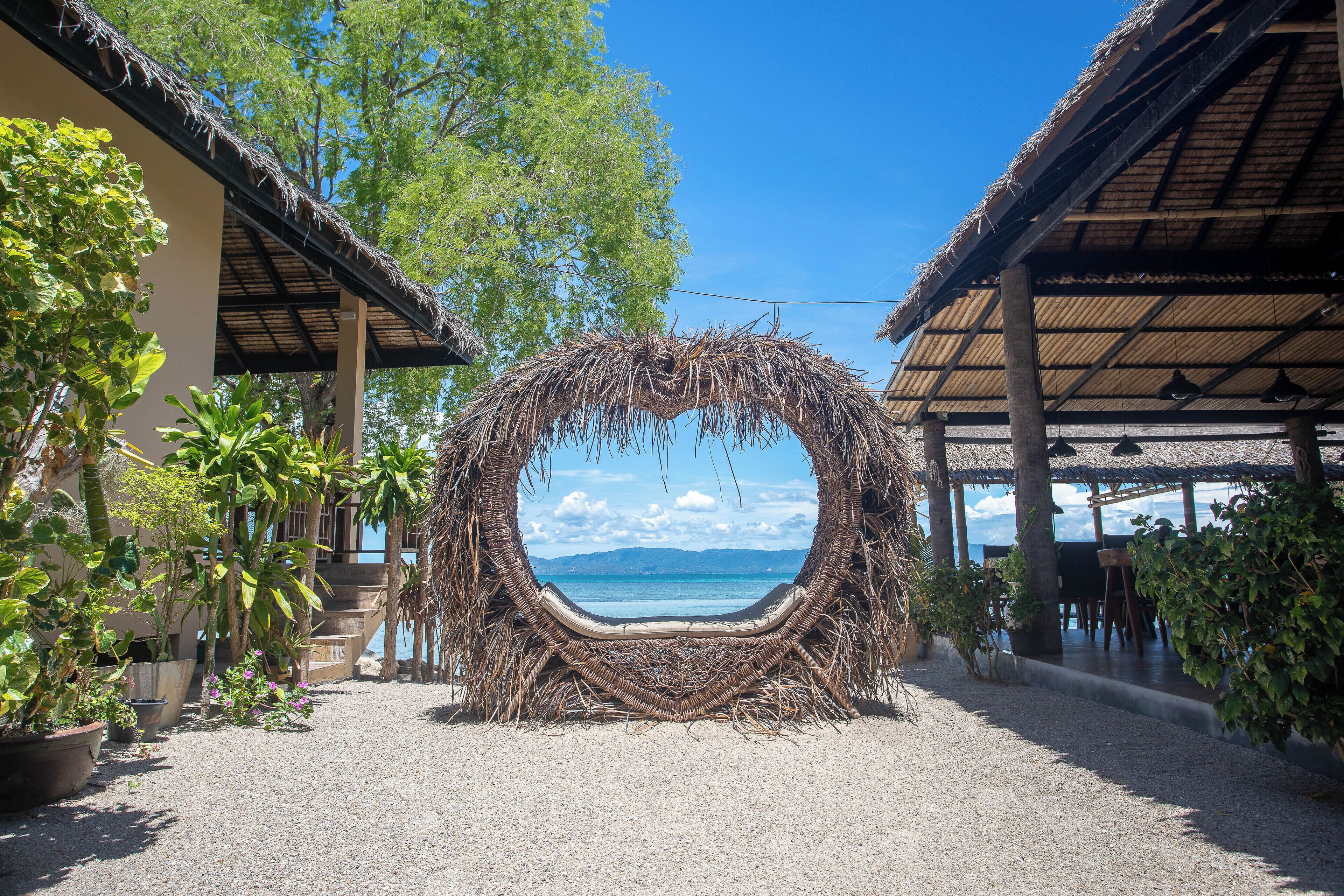 A circular wicker structure with a view of the sea, surrounded by potted plants and a covered seating area.