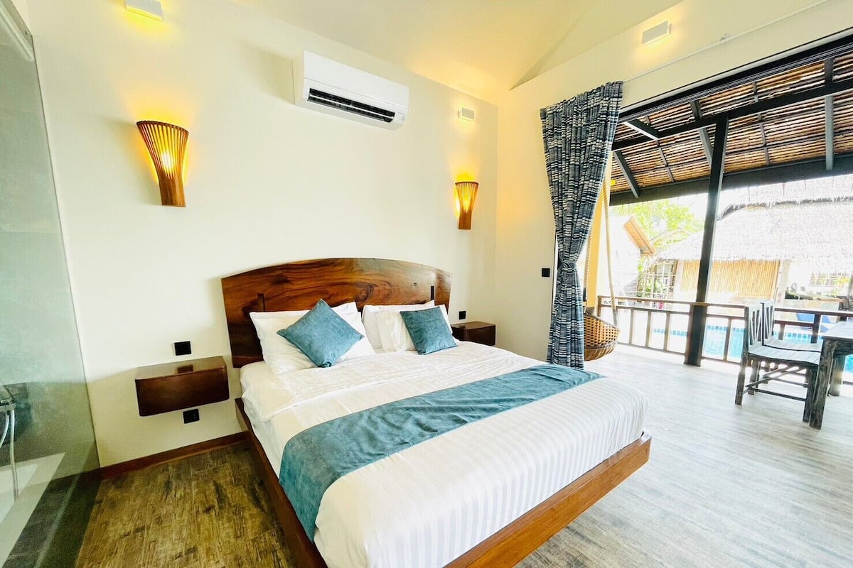 A bedroom with a large bed, two bedside tables, an air conditioning unit, and a view of an outdoor area with a table and chairs.