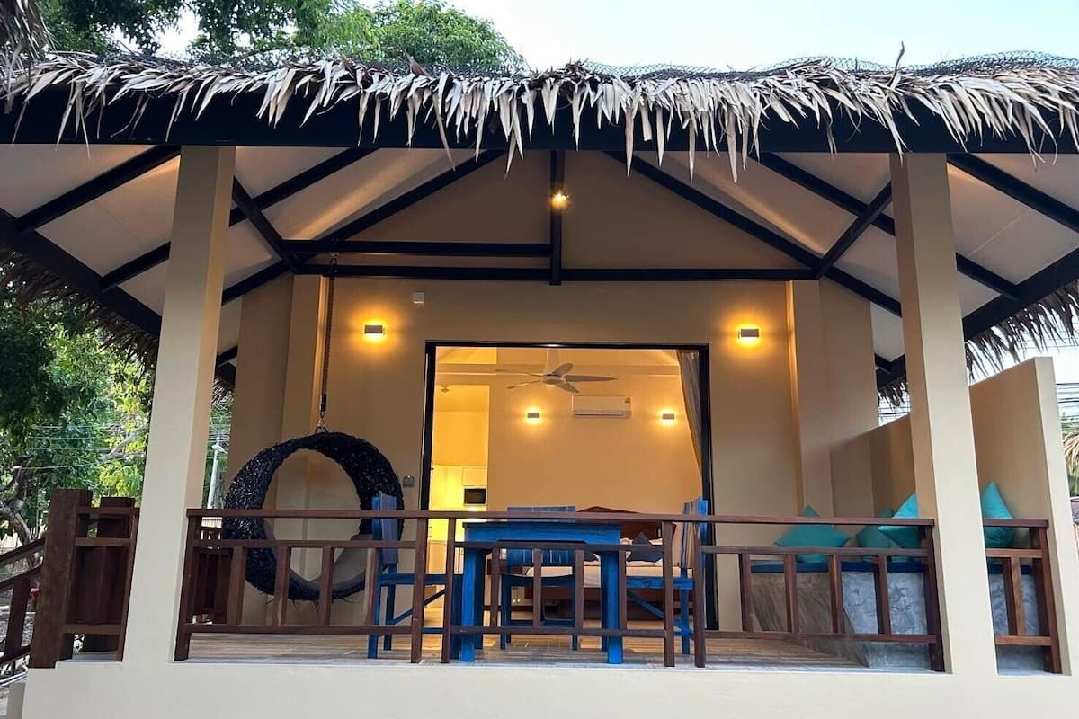 A covered outdoor seating area with a table and chairs, a thatched roof, and a view of the interior.