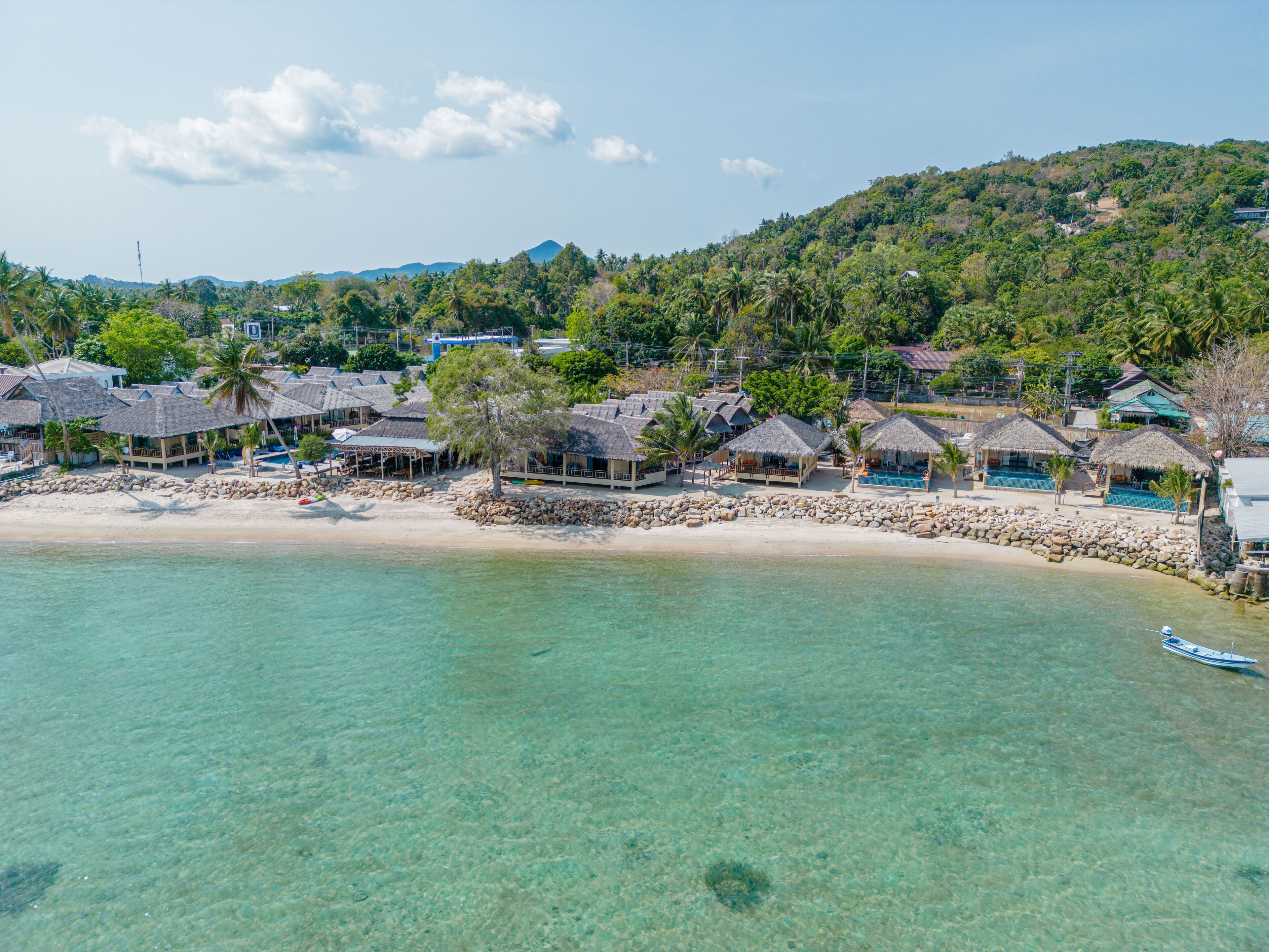 A coastal resort with thatched-roof bungalows, clear turquoise waters, and a rocky shoreline.