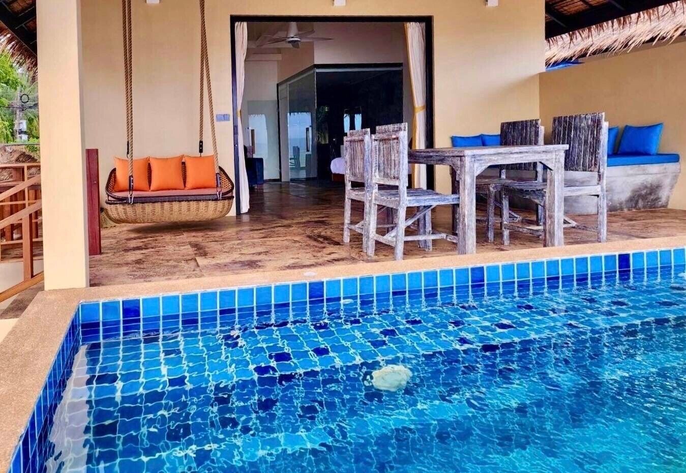 Un área junto a la piscina con una mesa y sillas de madera, un columpio de mimbre y una piscina con azulejos azules.