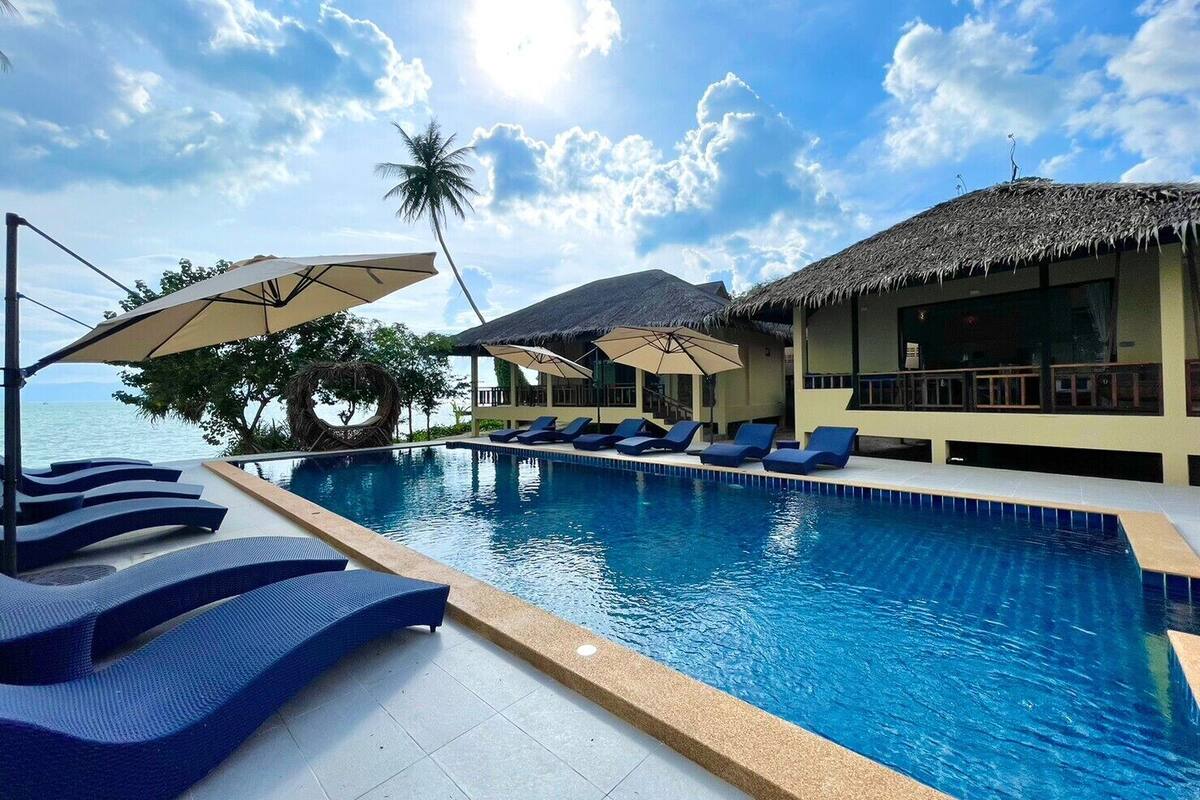 A poolside area with lounge chairs and umbrellas, a thatched-roof structure, and a view of the sea.