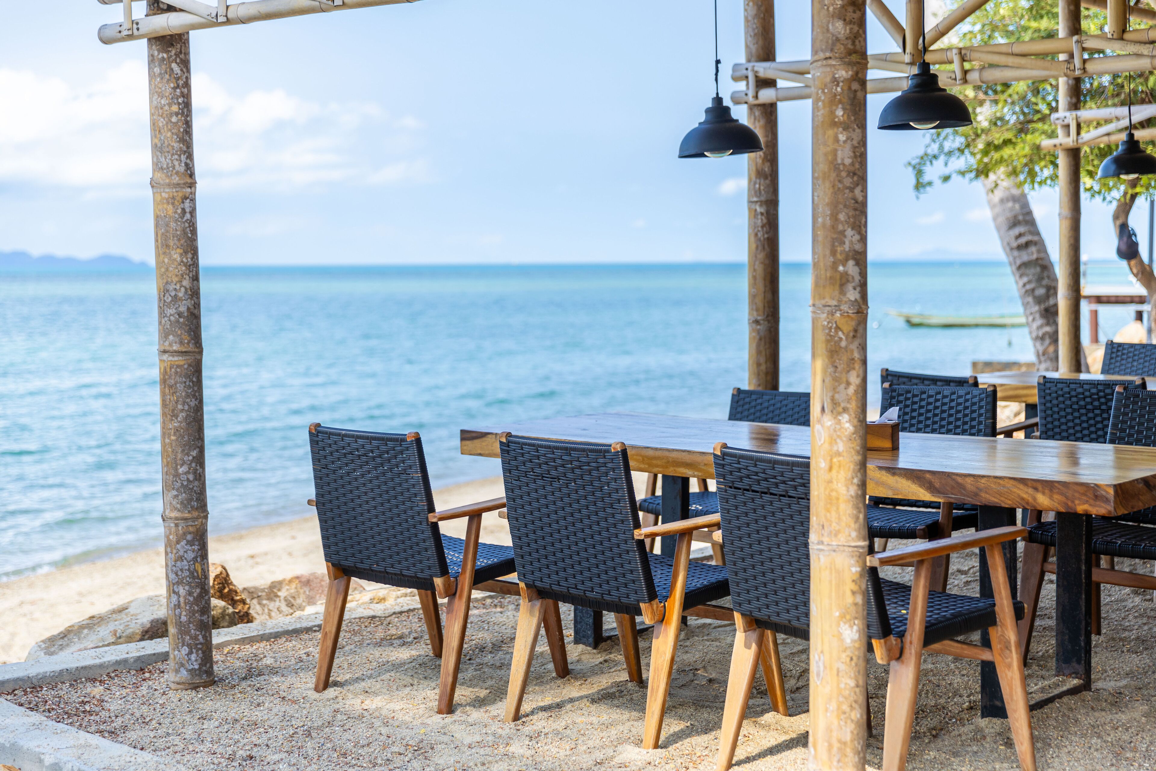 An outdoor dining area with a wooden table and chairs, overlooking a beach and the sea.