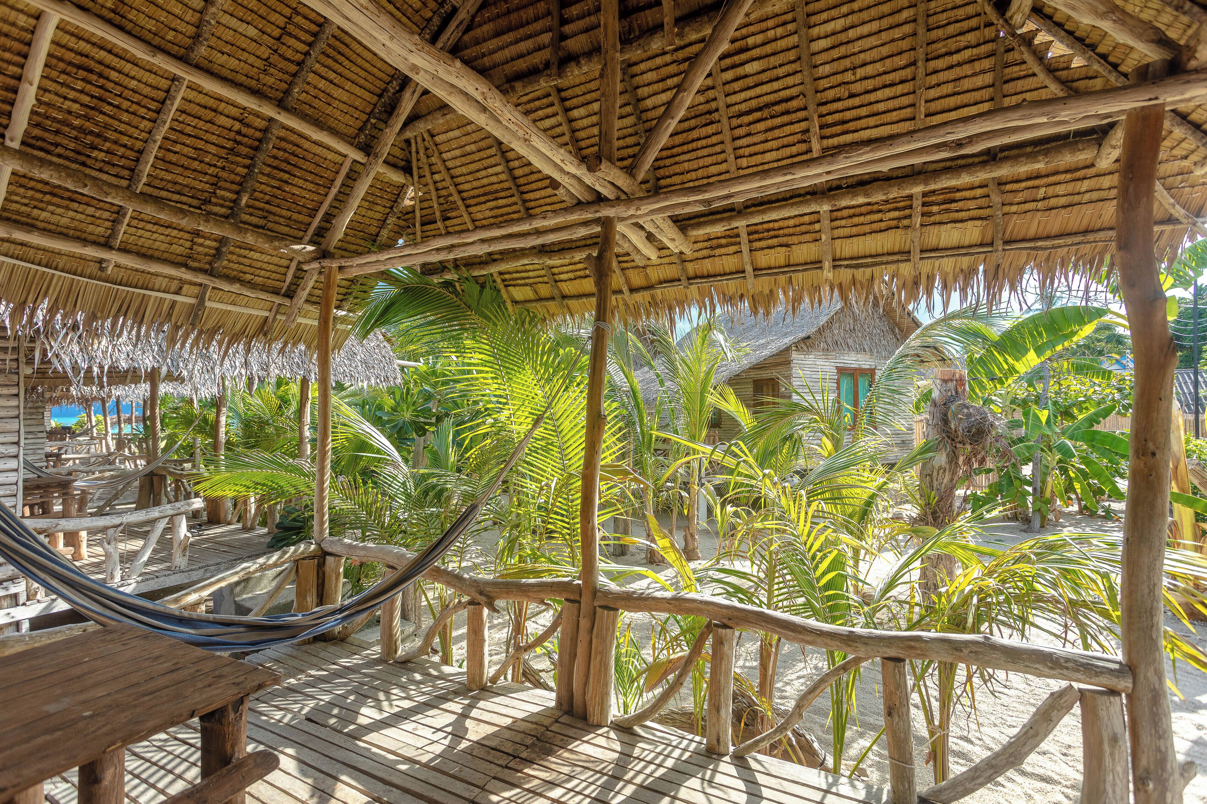 Une terrasse en bois surmontée d’un toit de chaume, entourée de palmiers et agrémentée d’un hamac.