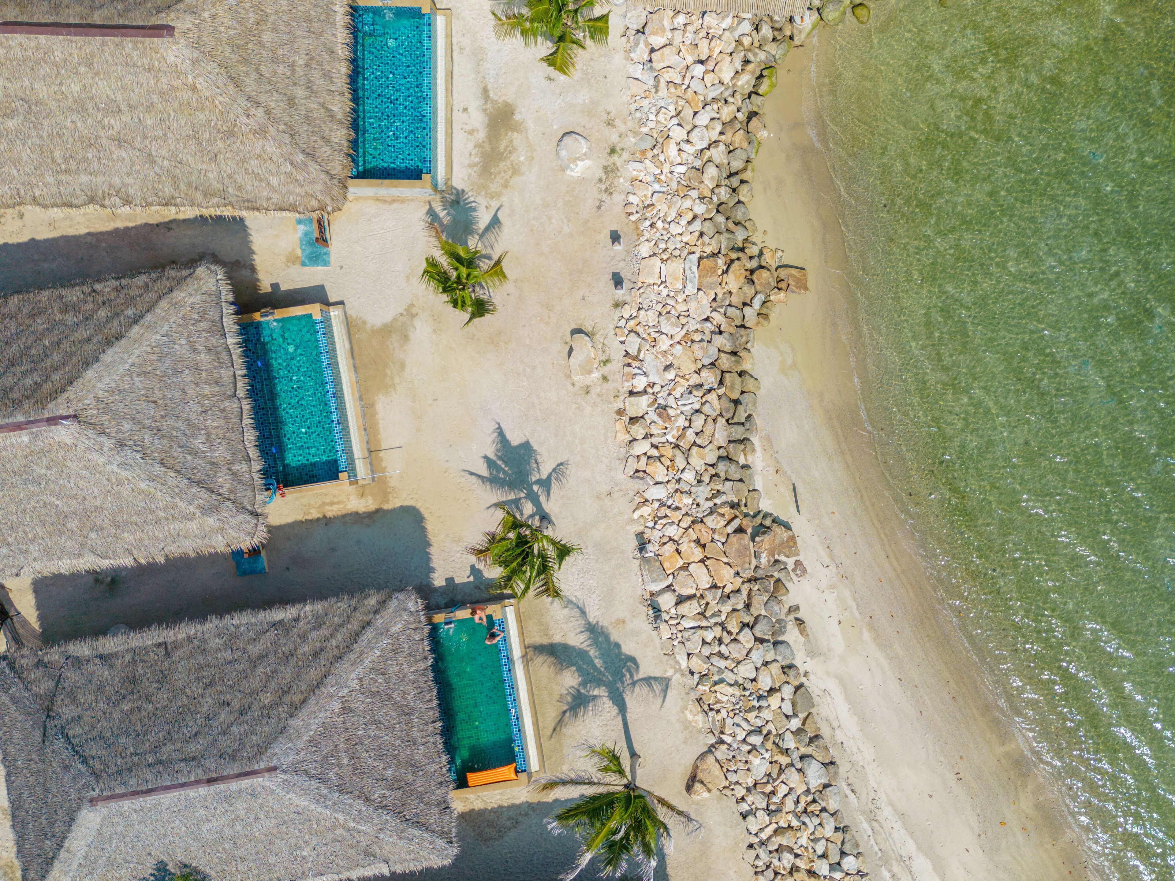 An aerial view of a beach resort with multiple swimming pools, a rocky shoreline, and palm trees.