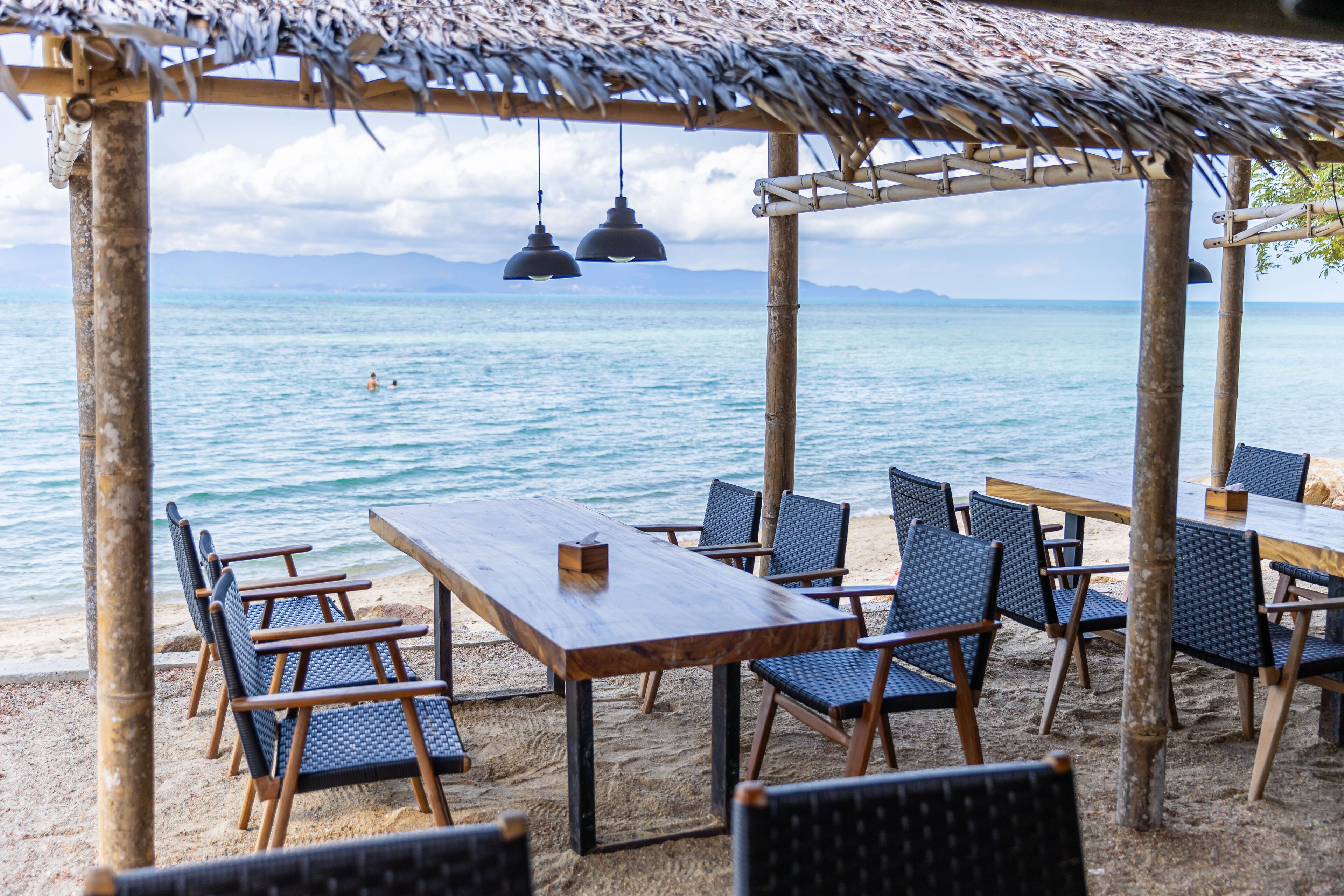 A beachside dining area with wooden tables and chairs, a thatched roof, and hanging lights.