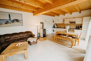 A cozy living room with a brown leather sofa, a wooden coffee table, and a dining area with a wooden table and chairs. A fireplace is visible in the background, and a kitchen area with white cabinets and appliances is to the right. - 'Haus Mimi Zarrentin' on Lake Schaalsee with Private Terrace & Garden (Zarrentin am Schaalsee)