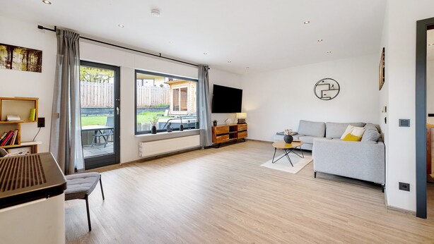 Een ruime woonkamer met een grijze bank, een flatscreen-tv aan de muur en een grote schuifpui die leidt naar een buitenruimte.