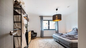 Een slaapkamer met een stapelbed, een slaapbank, een hanglamp, een raam met gordijnen en een radiator.