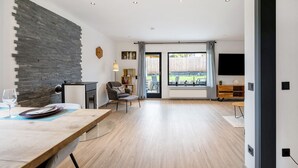 Een moderne woonkamer met een stenen muur, een eethoek met gedekte tafel, een bank en een aan de muur bevestigde televisie.