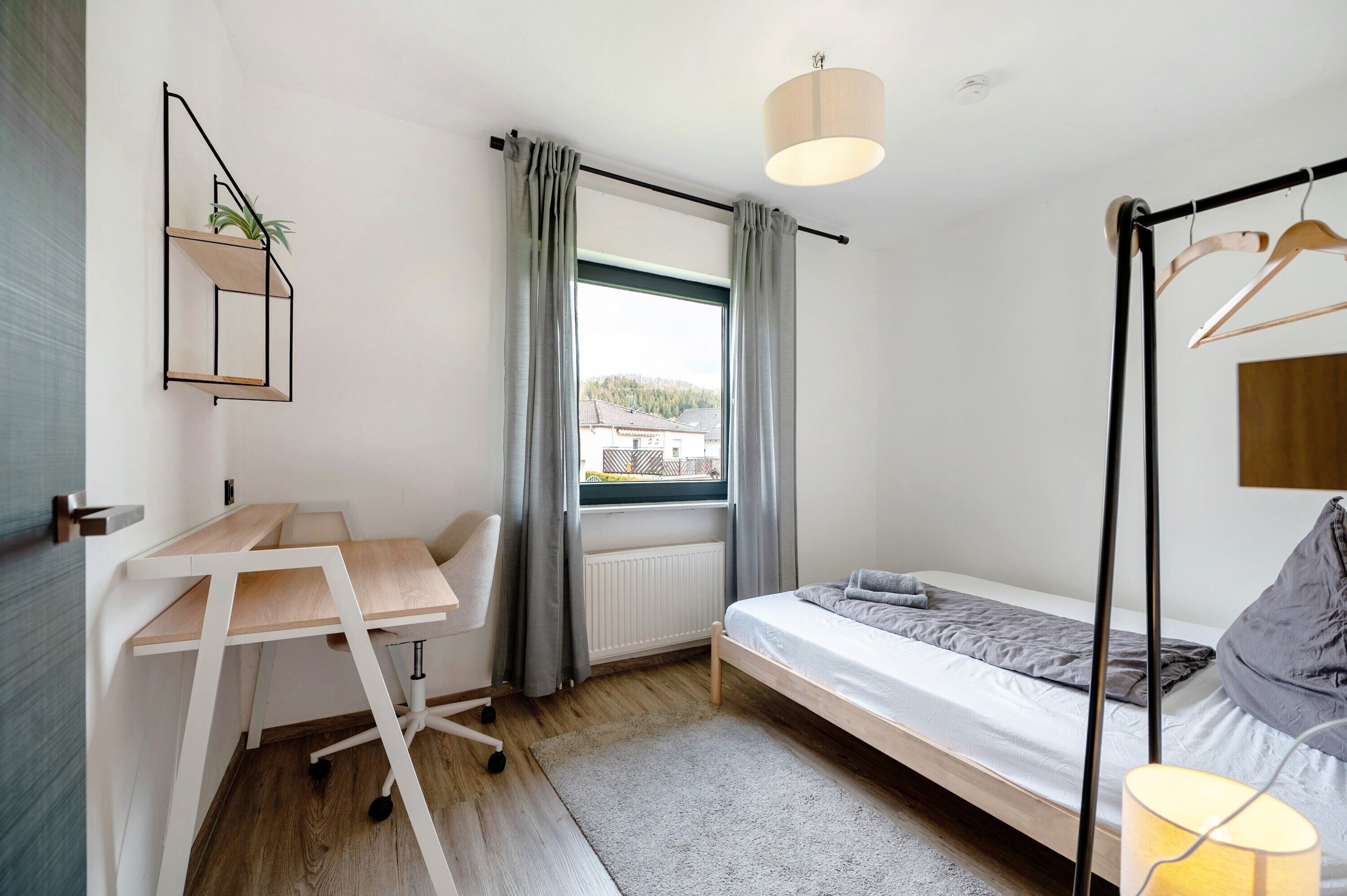 A compact bedroom with a bed, a desk, a chair, a radiator, and a window with curtains.