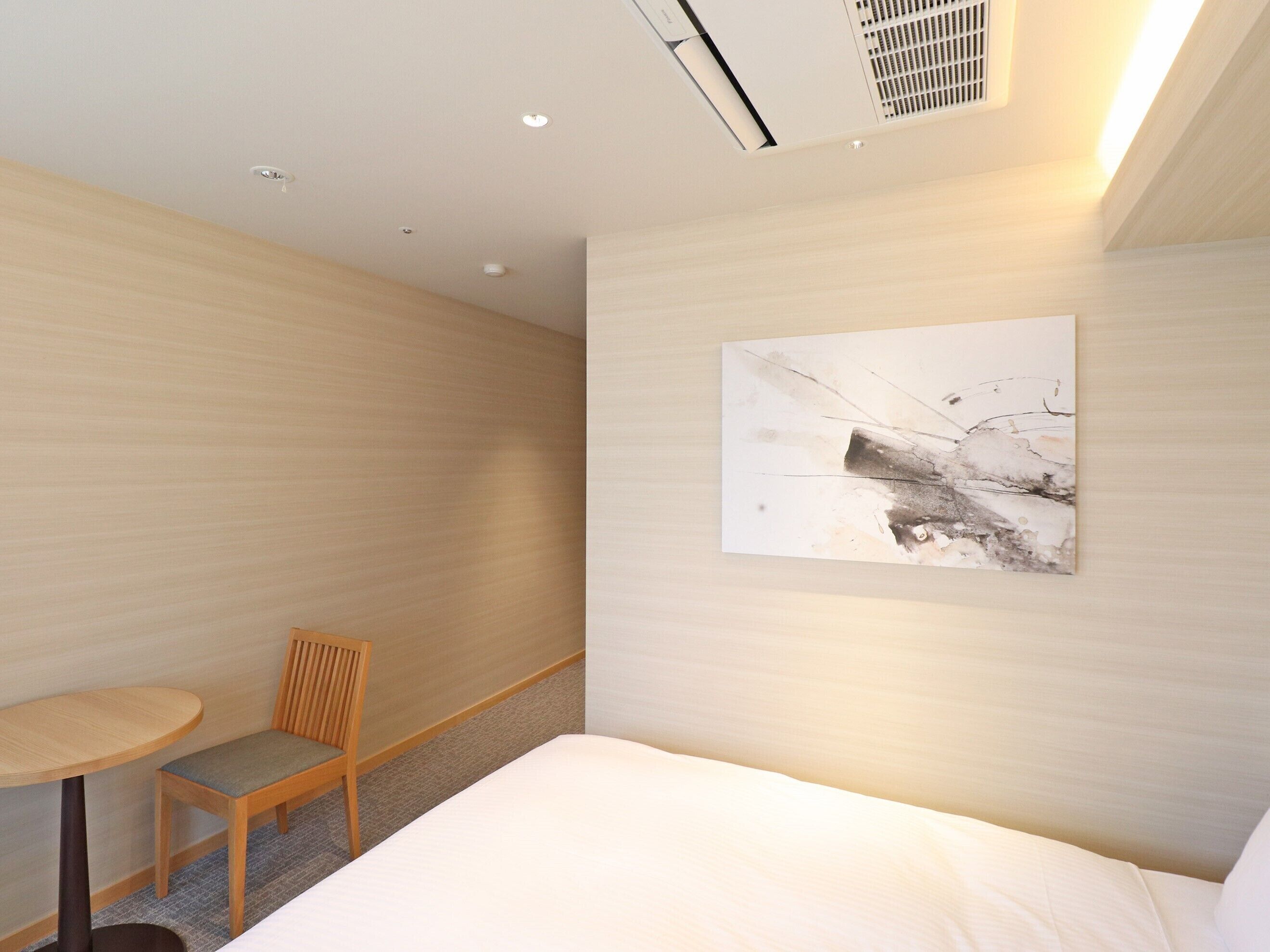 A modern hotel room with a large bed, a small wooden chair, a round table, and a wall-mounted abstract painting.