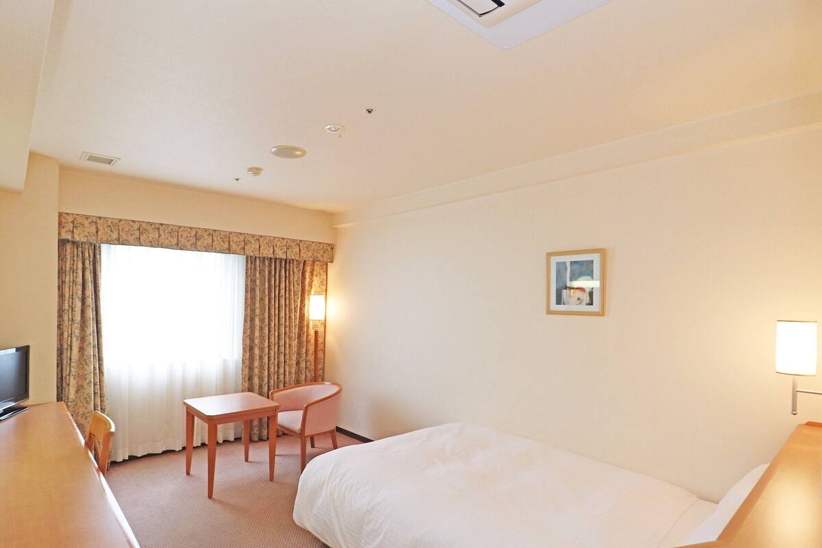 A hotel room with a bed, a desk, a chair, a TV, a lamp, and a window with curtains.
