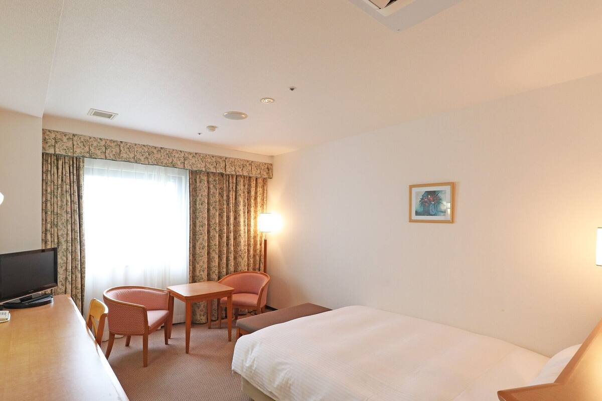 A hotel room with a bed, a desk with a TV, a chair, a small table, and a window with curtains.
