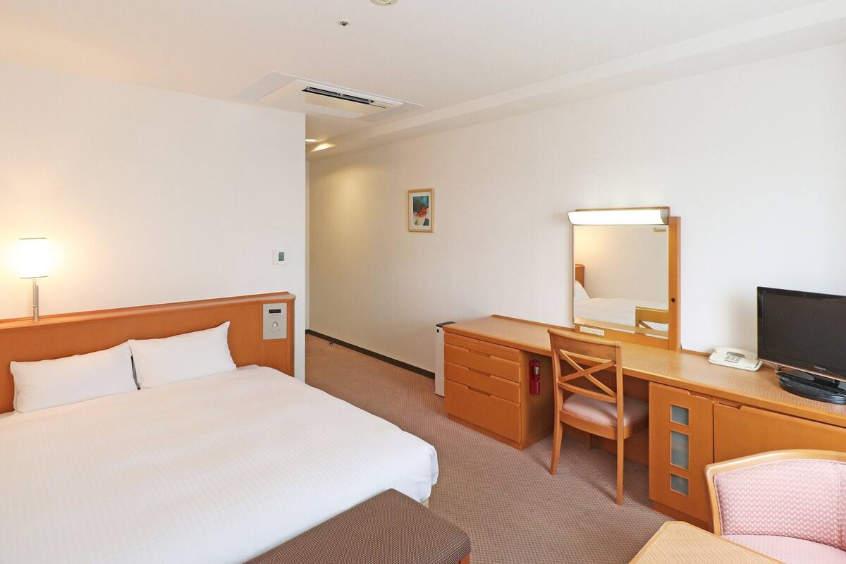 A hotel room with a bed, a desk with a chair, a television, and a bedside table.