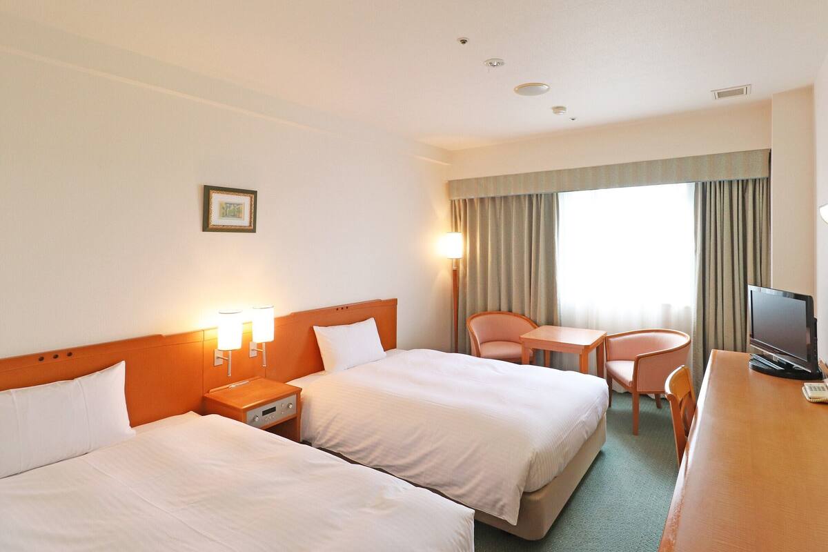 A hotel room with two beds, a desk, a chair, a TV, and a window with curtains.