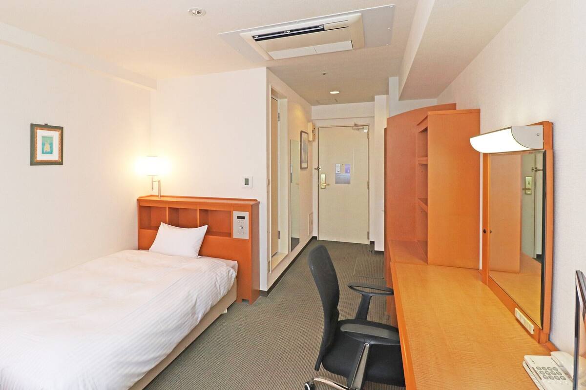 A hotel room with a single bed, a desk with a chair, a mirror, and an air conditioning unit.