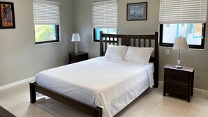 Une chambre à coucher comprenant un lit, deux tables de chevet, une commode et deux fenêtres équipées de stores.