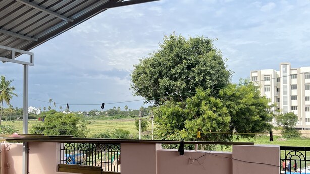 A covered outdoor area with a brick floor, a metal railing, and a view of a tree and buildings in the distance.