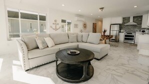 Una sala moderna con un sofá modular, una mesa de centro redonda y una cocina con armarios y electrodomésticos blancos.