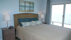A bedroom with a large bed, two bedside tables, a lamp, a remote control, and a view of the sea through a window.