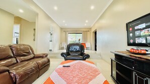 A living room with a brown leather sofa, a black armchair, a TV on the wall, and a dining area in the background.