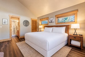 A bedroom with a large bed, wooden headboard, two bedside tables with lamps, a wooden door, and a wall with framed pictures.