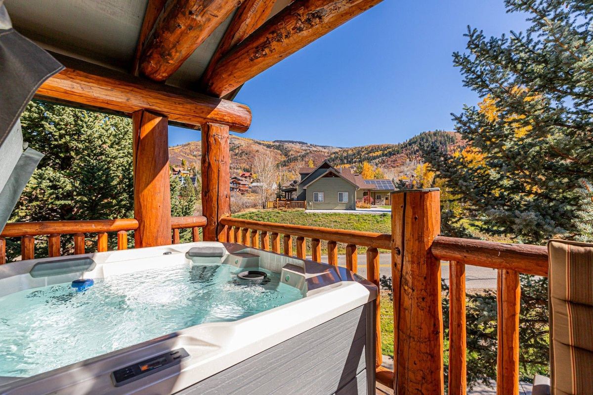 A hot tub on a wooden deck with a view of a mountain and a house in the background.